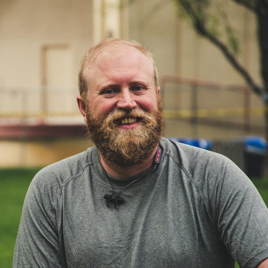 A cheerful bearded man smiling outdoors in a casual setting with greenery.